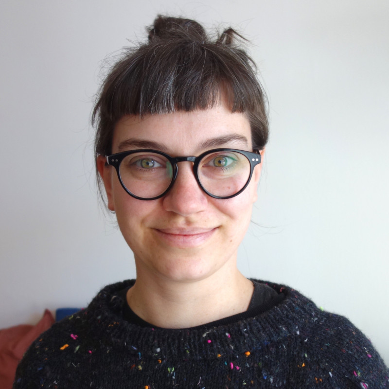 Photo d'une femme blanche souriante, avec lunettes et pull noir, cheveux bruns attachés avec frange