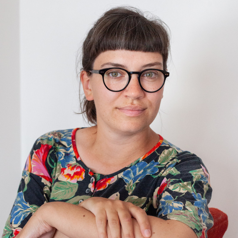 Photo d'une femme blanche souriante, avec lunettes et t-shirt coloré, cheveux bruns attachés avec frange